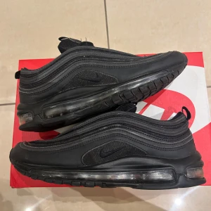 Nike Air Max 97 Triple Black sneakers - Nike Air Max 97 i helsvart färg med ikoniska vågformade linjer och synlig Air-sula runt hela skon. Ovandel i mesh och syntet, med svarta snören och diskret Swoosh-logga på sidan. Perfekta för dig som gillar en clean och sportig look. Finns slitage här och där men som syns väldigt lite, nypris ligger på 1600-2000kr. Pris kan diskuteras!😁