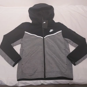 Nike hoodie svart och grå M - Nike hoodie i svart och grått med vit logga på bröstet. Tröjan har hel dragkedja, huva och långärmade ärmar. Snygg kontrast mellan färgerna och en sportig look. Perfekt för chill eller träning. Materialet är mjukt och känns bekvämt mot huden.