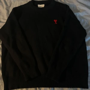 Svart stickad tröja från Ami - Svart stickad tröja från Ami med rund halsringning och ribbade muddar. På bröstet finns Ami:s ikoniska röda hjärta med bokstaven A broderat. Perfekt för dig som gillar stilrena och trendiga plagg med en twist.