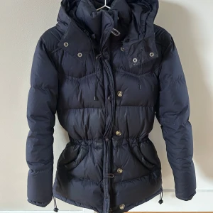 Mörkblå dunjacka med huva och snörning - Snygg mörkblå dunjacka med huva och dragsko i midjan för en schysst passform. Jackan har både knappar och dragkedja framtill samt stora fickor. Perfekt för kalla dagar när du vill hålla värmen och ändå se stilren ut.