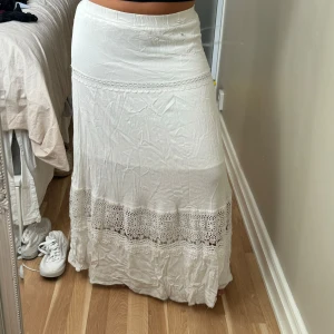Vit långkjol med spetsdetaljer - Romantisk vit långkjol med breda spetsinslag nertill och dekorativa sömmar. Kjolen har en flowy passform och är tillverkad i ett lätt tyg som känns perfekt för varma dagar. Den har en bohemisk vibe och passar dig som gillar en avslappnad men söt stil. Tyvärr har en gått sönder lite men ingen man lägger märke till om man inte kollar nära