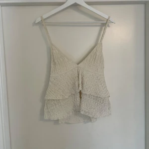 Beige volangig linnetopp - Säljer en söt beige linnetopp med volanger och smala axelband. Toppen har en lätt skrynklig struktur och v-ringad halsringning både fram och bak. Perfekt för varma dagar och enkel att matcha med jeans eller kjol.