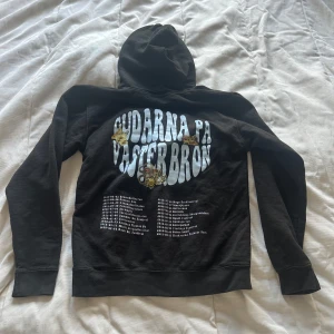 Svart Gudarnas Hoodie Västerbron - Svart hoodie med stor vit tryckt text 'Gudarnas på Västerbron' och festivalturné på ryggen. Framsidan har en liten broderad krona på bröstet. Klassisk känguruficka, snörning i huvan och mjukt bomullsmaterial. Perfekt för dig som gillar hov1.💕 är en storlek S men skulle säga mer som xs.