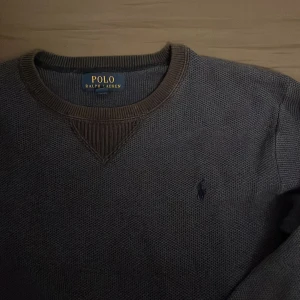 Blå stickad tröja från Polo Ralph Lauren - Mörkgrå stickad tröja från Polo Ralph Lauren med klassisk rund halsringning och diskret broderad logga på bröstet. Tröjan har ribbade muddar och en snygg detalj vid halsen. Perfekt för lager-på-lager under kyliga dagar.