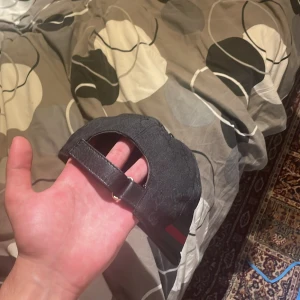 Svart Gucci keps med GG-mönster - Snygg svart keps från Gucci med klassiskt GG-monogram över hela kepsen. Kepsen har justerbart spänne bak och en ikonisk grön-röd rand på sidan. Materialet är bomull och kepsen har en böjd skärm för en sportig look.