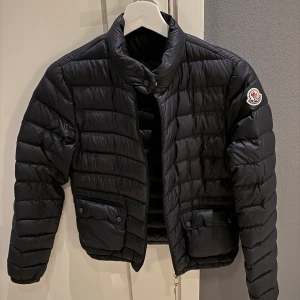 Svart dunjacka från Moncler modell: LANS GIUBBOTTO LANS JACKET - Säljer en svart dunjacka från Moncler med quiltad design och hög krage. Klassisk Moncler-logga på ena ärmen. Väldigt varm men andningsbar så går att använde både i höst och vinter! Skriv privat om ni vill ha fler bilder eller verifiera att den är äkta💕minns tyvärr inte vilken storlek men skulle gissa på storlek 0/xs!