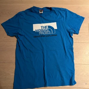 Blå t-shirt från The North Face - Snygg blå t-shirt från The North Face med stor vit logga och texten 'Never Stop Exploring' på bröstet. Klassisk rund hals och korta ärmar. Tillverkad i mjuk bomull som är skön mot huden. Perfekt för dig som gillar streetwear och outdoor-vibe.