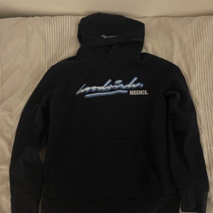 Svart hoodie från Hoodrich med blått tryck - Svart hoodie från Hoodrich med stor blå och vit logga framtill. Klassisk känguruficka, huva och ribbade muddar. Snygg streetstil med tryck även på ryggen. Perfekt för dig som gillar urban vibe och vill sticka ut lite extra.