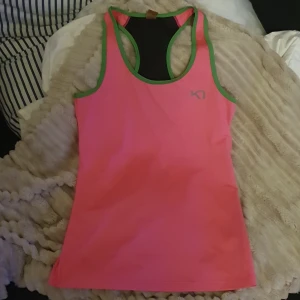 Kari Traa rosa träningstopp. S - Obs, små prickar från hårfärg på baksidan vid nacke/ axlar. Syns på foto.                        Sportig rosa linnetopp från Kari Traa med gröna kanter och svart meshpanel i ryggen. Perfekt för träning och har en tight passform som sitter snyggt. Snygg logga framtill och bak. Materialet är syntet för bästa komfort under aktivitet.