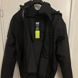 Svart vindjacka från Helly Hansen - Svart vindjacka från Helly Hansen. Storlek L men passar mer M. 