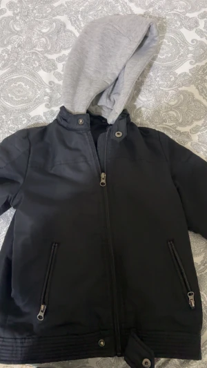 Svart bomberjacka med grå hoodie från Ellos - Svart bomberjacka från Ellos med grå huva i sweatshirt-tyg. Jackan har dragkedja framtill, två fickor med dragkedja och snygga detaljer vid kragen och nederkanten. Perfekt för dig som gillar streetstyle och vill ha en cool look.