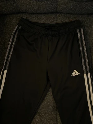 Svarta Adidas träningsbyxor - Svarta träningsbyxor från Adidas med  vita och grå ränder längs sidorna. Byxorna har elastisk midja och sidofickor, samt Adidas-logga på låret. Passar 11-12Y.
