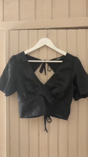 Svart omlottopp från Bik Bok - Snygg svart omlottopp från Bik Bok med croppad passform. Toppen har korta puffärmar, v-ringning och knytband både i ryggen och nedtill. Materialet är glansigt och känns lyxigt, perfekt för att lyfta din outfit.