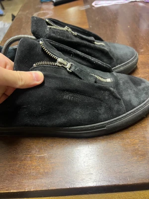 Axel Arigato Svarta sneakers med dragkedja - Svarta sneakers från Axel Arigato i mocka med coola silverfärgade dragkedjor framtill. Handgjorda i Portugal, har rund tå och platt sula. Baksidan är i svart skinn för extra stilpoäng. Perfekt för dig som gillar minimalistisk design med en twist.