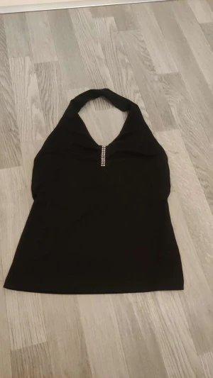 Svart halterneck topp med strass - Svart halterneck topp från Hennes i storlek 36/38. Toppen har en snygg detalj med strass framtill vid urringningen och är ärmlös med en tight passform. Perfekt för dig som gillar stilrena plagg med lite extra bling.