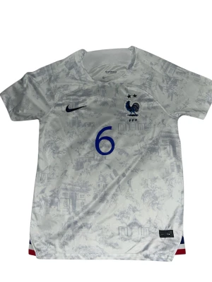 Frankrike Pogba #6 Nike fotbollströja - Snygg vit Frankrike fotbollströja från Nike med Pogba och nummer 6 i blått på ryggen. Tröjan har korta ärmar, diskret mönster med franska motiv och FFF-logga med tupp på bröstet. Materialet är lätt och andas, perfekt för match eller träning.