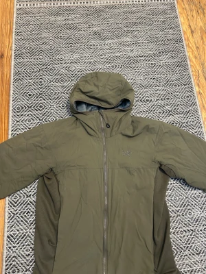 Grön vindjacka från Arc'teryx - Säljer en snygg grön vindjacka från Arc'teryx med huva och hel dragkedja framtill. Jackan har ett stilrent Arc'teryx-logo broderat på bröstet och är gjord i lätt polyester. Perfekt för dig som gillar outdoor och streetwear.