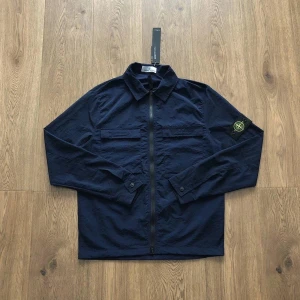 Stone Island Overshirt - Snygg mörkblå overshirt från Stone Island med klassisk logotyp-patch på ärmen. Modellen har två bröstfickor med lock, hel dragkedja framtill och krage. Materialet är lätt och känns skönt mot huden, perfekt för lager-på-lager. Långa ärmar med knapp vid manschetten.