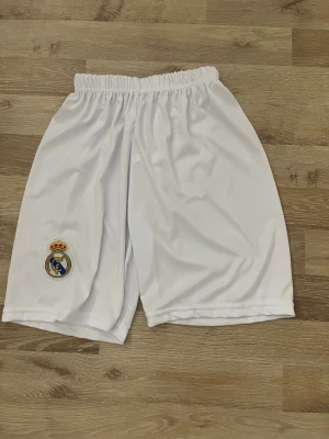 Vita Real Madrid fotbollsshorts - Säljer ett par vita fotbollsshorts från Real Madrid med klubbens broderade emblem på vänster ben. Shortsen har elastisk midja och är gjorda i ett lätt och skönt polyestermaterial, perfekta för träning eller match. Klassisk och sportig look för alla fans.