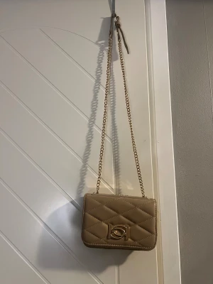 Beige/brun quiltad axelväska med kedja - Snygg beige/brun axelväska med quiltad design och guldfärgad kedja som axelrem. Väskan har en stilren rektangulär form och en elegant detalj framtill. 