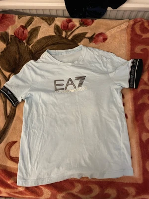 Ljusblå EA7 Emporio Armani t-shirt - Snygg ljusblå t-shirt från EA7 Emporio Armani med stort logotryck på bröstet och svarta detaljer med logga på ärmsluten. Klassisk rund hals och rak passform. Perfekt för dig som gillar stilrena märkesplagg.