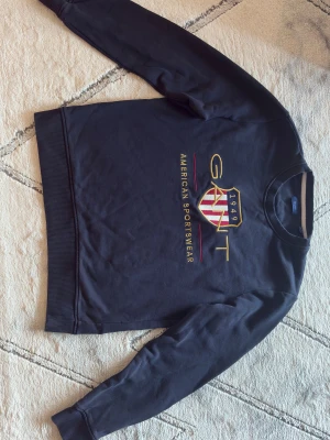 Mörkblå sweatshirt från GANT - Snygg mörkblå sweatshirt från GANT med broderad logga och emblem i guld, rött och vitt. Tröjan har rund hals, ribbade muddar och lång ärm. Perfekt för dig som gillar klassisk amerikansk sportstil. str M/38