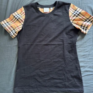 Svart Burberry t-shirt med rutiga ärmar - Svart t-shirt från Burberry med klassiska rutiga korta ärmar i beige, svart, vit och röd. Rund halsringning och normal passform. Materialet är mjuk bomull och ärmarna har det ikoniska Burberry-mönstret som ger en lyxig vibe. Perfekt för dig som gillar stilrena plagg med en twist.