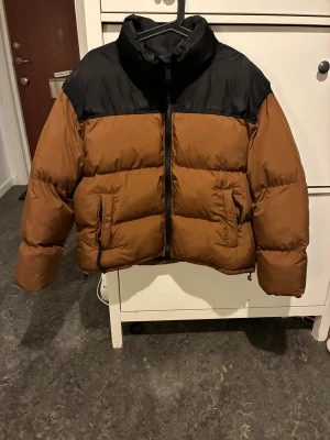 Brun och svart pufferjacka Authorized - Säljer en oversized pufferjacka från Authorized i brun och svart. Jackan har hög krage med broderad logga, dragkedja framtill och två fickor med dragkedja. Materialet är syntet och jackan har en riktigt puffig look som är perfekt för kalla dagar.