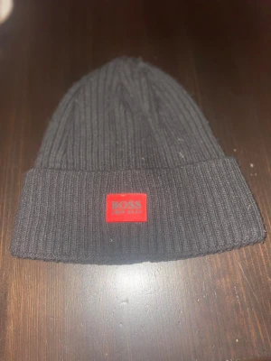 Svart ribbad mössa från Hugo Boss - Svart ribbstickad mössa från Hugo Boss med uppvikt kant och röd patch framtill där det står BOSS. Klassisk och stilren design som passar perfekt till kalla dagar. Tillverkad i mjukt material för extra komfort.