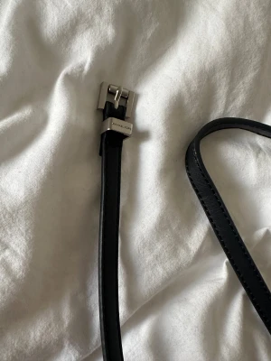 Svart smalt skärp från Michael Kors - Snyggt svart skärp från Michael Kors med smal design och stilren silverfärgad metallspänne. Skärpet är gjort i slätt skinn och har diskreta sömmar längs kanterna. Perfekt för att ge din outfit en clean och modern touch. Storlek 4 Längd: ca 95cm