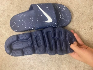 Blå Nike sandaler med vita stänk - Säljer ett par blå Nike sandaler med coolt vitt stänkmönster och stor vit Swoosh-logga. De har en bred rem över foten och en mjuk, formad sula för extra komfort. Perfekta för sommaren eller att chilla i hemma.