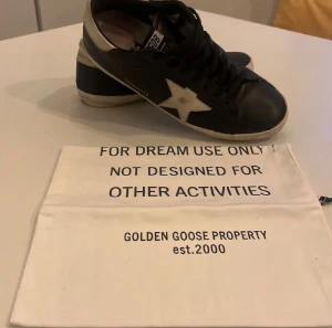 Golden Goose Skor - Golden Goose sneakers i svart skinn med vit stjärndetalj på sidan och vit kant vid hälen. Klassisk låg modell med snörning och platt sula. Snyggt sliten look och GGDB/SSTAR-tryck på sidan. Kommer med originalpåse och certifikat. 2 små defekter vid hälarna dock inget som påverkar skon och inget som syns när du använder skorna. Priset är ej hugget i sten och är också öppen mot byten 