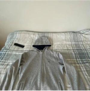 Grå hoodie från Lacoste med dragkedja - Säljer en stilren grå hoodie från Lacoste med hel dragkedja och huva. Tröjan har långa ärmar och den klassiska Lacoste-loggan på bröstet. Perfekt för chill dagar och enkel att matcha med jeans eller joggers.