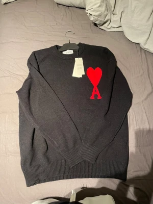 Svart stickad tröja från Ami Paris - Svart stickad tröja från Ami Paris med röd hjärtapplikation och bokstaven A på bröstet. Rund hals, ribbade muddar och långärmad. Tröjan är tillverkad i Portugal och har en clean, klassisk look med snygg detalj framtill.