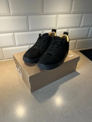 Christian Louboutin svarta sneakers - Snygga svarta sneakers från Christian Louboutin med klassisk röd sula och svarta skosnören. Ovandel i mocka och insida i ljust skinn. Kommer med originalkartong, dustbag och papper. Perfekta för dig som vill ha en lyxig och stilren look. Sparsamt använda 