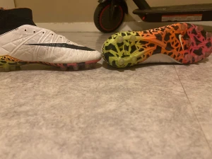 Nike fotbollsskor med färgglad sula - Säljer ett par Nike fotbollsskor med hög strumpkrage i svart och vit ovandel. Skorna har en cool sula med mönster i neonfärger som gul, orange och röd, samt svarta detaljer. Ovandelen är tillverkad i syntetmaterial och har en rund tå. Perfekt för dig som vill sticka ut på planen.