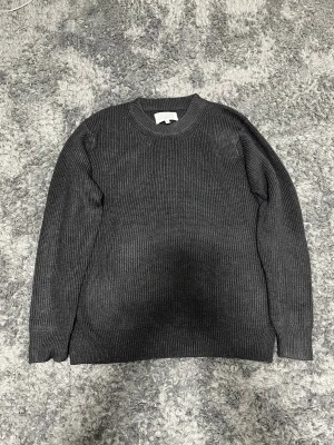 Mörkgrå stickad tröja från Maison Margiela - Snygg mörkgrå stickad tröja från Maison Margiela med klassisk rund hals och ribbade muddar. Tröjan har långärmad passform och diskreta vita stygn i nacken som är typiska för märket. Perfekt för dig som gillar stilrena och tidlösa plagg.