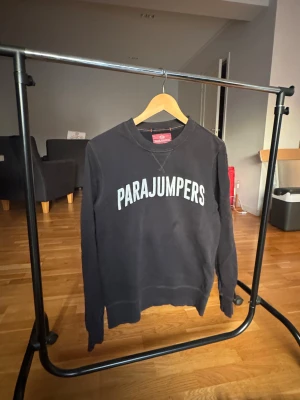 Svart fleece-tröja Parajumpers S - Svart fleece-tröja från Parajumpers i storlek S med vit logotyp på bröstet och snyggt broderat märke på ärmen. Tröjan har även orangea sömmar. Perfekt för dig som gillar streetwear och vill ha något bekvämt. Har använts sparsamt med inga defekter.