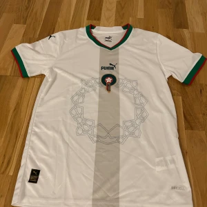 Vit Marocko fotbollströja Puma S - Säljer en officiell Marocko fotbollströja från Puma i storlek S. Tröjan är vit med subtilt geometriskt mönster på framsidan, gröna och röda detaljer vid krage och ärmslut samt svarta Puma-loggor på axlarna. Tillverkad i lätt och ventilerande polyester med DryCell-teknologi.