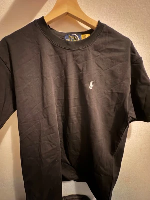 Svart t-shirt från Polo Ralph Lauren - Svart t-shirt från Polo Ralph Lauren med klassisk passform och rund hals. Ikoniska vita broderade loggan på bröstet. Tillverkad i mjuk bomull som känns skön mot huden. Perfekt basic-plagg med stilren look.