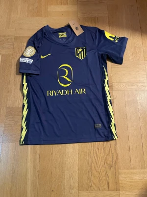 Atlético Madrid x Nike blå matchtröja - Snygg blå Atlético Madrid fotbollströja från Nike med gula detaljer och tryck, inklusive klubbmärke och sponsorlogga. Kortärmad modell i lätt och ventilerande material, perfekt för match eller träning. Blixtdetaljer längs sidorna och officiella FIFA Club World Cup 2025-märken på ärmen.