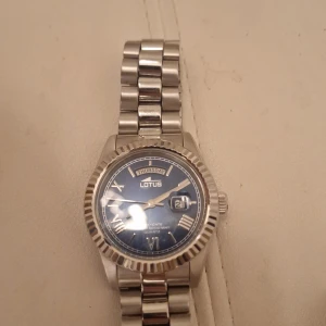 Silverfärgad Lotus klocka med blå urtavla - Stilren klocka från Lotus med silverfärgat metallarmband och blå urtavla. Urtavlan har romerska siffror, dag- och datumvisning samt markerad ram runt glaset. Perfekt accessoar för dig som gillar klassisk design med modern touch.