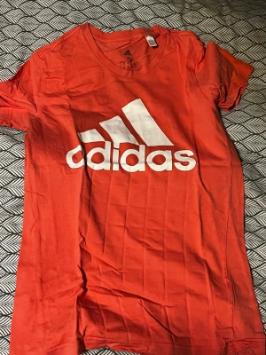 Röd Adidas t-shirt med vit logga - Snygg röd t-shirt från Adidas med stor vit logga på bröstet. Klassisk rund hals och korta ärmar. Tillverkad i mjuk bomull som är skön mot huden. Perfekt för dig som gillar sportig stil och vill sticka ut med en färgstark look.