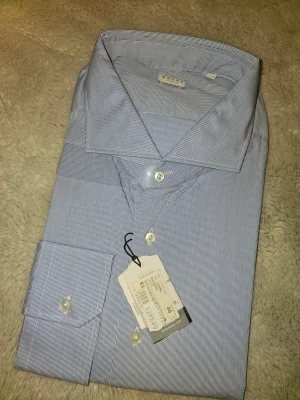 Blårandig skjorta från Xacus, strl 46 - Snygg skjorta från Xacus i tailor fit med tunna blå och vita ränder. Klassisk krage, vita knappar och lång ärm. Skjortan är gjord i mjuk bomull och har en stilren look som passar perfekt till jeans eller chinos. Märkt med storlek 46.