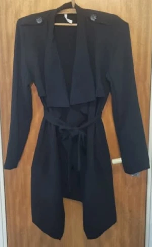 Svart trenchcoat med bälte - Snygg svart trenchcoat med långa ärmar och bred krage. Jackan har axelklaffar med dekorativa knappar och ett knytbälte i midjan för att skapa en schysst siluett. Perfekt för dig som gillar stilrena och klassiska plagg.