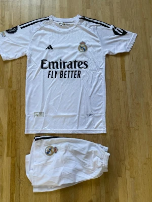 Real Madrid Mbappé 9 fotbollströja Adidas - Säljer en vit Real Madrid fotbollströja från Adidas med Mbappé och nummer 9 på ryggen. Tröjan har korta ärmar, svarta detaljer och klubbmärke på bröstet. Materialet är lätt och andas, perfekt för match eller träning. Shorts med klubbmärke ingår.