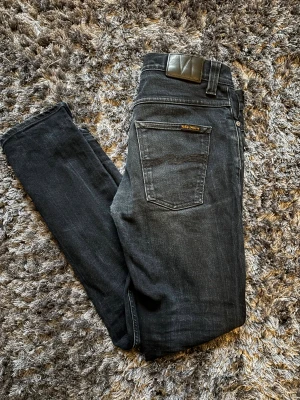 Feta jeans från Nudie  - Säljer ett par svarta jeans från Nudie Jeans i modellen grim tim. De har klassisk femficksdesign, svarta sömmar och en snygg läderpatch bak i midjan. Jeansen är tillverkade i stretchig denim för extra komfort och har en smal passform hela vägen ner. Jeansen är i bra skick med snygga fades men har en lagning i skrevet som inte syns när jeansen är på.