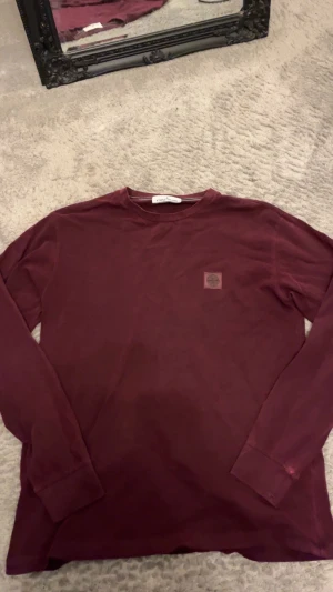 Stilren vinröd långärmad tröja Stone Island  - Snygg vinröd långärmad tröja från Stone Island med klassisk rund hals och ikonisk patch på bröstet. Tröjan är i mjuk bomull och har en clean look som passar perfekt till jeans eller cargo. Perfekt för dig som gillar streetwear och vill ha något stilrent.