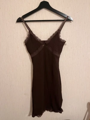 Brun ribbad spetsklänning med rosett - Supersöt brun kortklänning med ribbad struktur och tunna axelband. Klänningen har spetsdetaljer längs urringning och fåll, samt en liten rosett framtill. Figurnära passform och stretchigt material som känns bekvämt och följsamt.