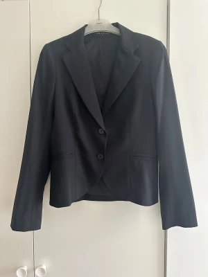 Svart kavaj från Theory - Snygg svart kavaj från Theory. Klassisk modell med två knappar framtill, spetsiga slag och med fickor. Perfekt för dig som gillar stilrena och tidlösa plagg. Kavajen är i storlek 8
